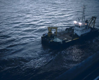 A small industrial vessel is navigating through slightly choppy waters. The vessel is equipped with machinery, including a yellow crane and antennas on its deck. The surrounding water has a dark, moody appearance due to the overcast lighting.