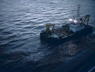 A small industrial vessel is navigating through slightly choppy waters. The vessel is equipped with machinery, including a yellow crane and antennas on its deck. The surrounding water has a dark, moody appearance due to the overcast lighting.