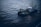 A small industrial vessel is navigating through slightly choppy waters. The vessel is equipped with machinery, including a yellow crane and antennas on its deck. The surrounding water has a dark, moody appearance due to the overcast lighting.
