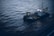 A small industrial vessel is navigating through slightly choppy waters. The vessel is equipped with machinery, including a yellow crane and antennas on its deck. The surrounding water has a dark, moody appearance due to the overcast lighting.