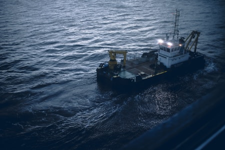 A small industrial vessel is navigating through slightly choppy waters. The vessel is equipped with machinery, including a yellow crane and antennas on its deck. The surrounding water has a dark, moody appearance due to the overcast lighting.