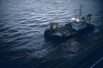 A small industrial vessel is navigating through slightly choppy waters. The vessel is equipped with machinery, including a yellow crane and antennas on its deck. The surrounding water has a dark, moody appearance due to the overcast lighting.