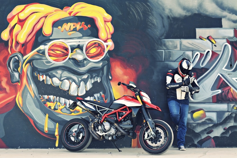 man in black jacket and blue denim jeans standing beside black and yellow motorcycle