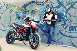 A person wearing full motorcycle gear, including a helmet and gloves, leans against a graffiti-covered wall featuring a large, artistic face. Next to them is a sporty red and white motorcycle. The graffiti is an elaborate design with abstract elements in blue, yellow, and black.
