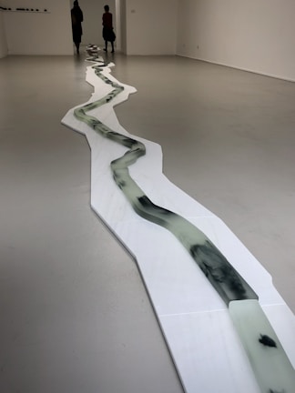 A minimalist art installation in a gallery setting features a long, winding structure resembling a river or path. The structure is composed of a series of connected panels with dark, fluid-like patterns. Two people are seen at the far end of the room, adding a sense of scale to the piece.
