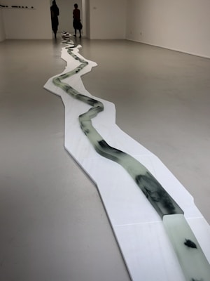 A minimalist art installation in a gallery setting features a long, winding structure resembling a river or path. The structure is composed of a series of connected panels with dark, fluid-like patterns. Two people are seen at the far end of the room, adding a sense of scale to the piece.