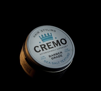 A round tin container prominently labeled 'CREMO' with additional text describing it as 'Barber Grade' and 'Sea Salt Texture.' The lid features a simplistic graphic of a crown and lists characteristics such as 'Low Hold' and 'Low Shine.' The container is partially illuminated against a dark background, creating a dramatic and focused presentation.