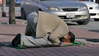 A man is kneeling on a mat on the sidewalk, appearing to be in prayer. The setting is an urban area with cars parked nearby on a sunny day.