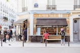 A quaint bakery with a traditional French façade occupies a street corner in a bustling city. People are walking by, and some are standing near the bakery, one person seated on a bench outside. The bakery window displays a variety of baked goods. Bicycles are parked nearby, and there's a small bird on the pavement, adding liveliness to the scene.