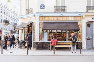 A quaint bakery with a traditional French façade occupies a street corner in a bustling city. People are walking by, and some are standing near the bakery, one person seated on a bench outside. The bakery window displays a variety of baked goods. Bicycles are parked nearby, and there's a small bird on the pavement, adding liveliness to the scene.