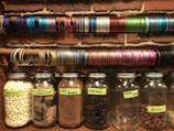 Several glass jars filled with various ingredients such as white chocolate chips, dark chocolate chunks, cocoa powder, hibiscus, cocoa nibs, and pecans are lined up on a shelf against a brick wall. Above the jars, there are multiple rows of colorful bangles arranged on rods.