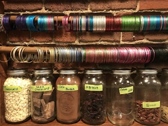 Several glass jars filled with various ingredients such as white chocolate chips, dark chocolate chunks, cocoa powder, hibiscus, cocoa nibs, and pecans are lined up on a shelf against a brick wall. Above the jars, there are multiple rows of colorful bangles arranged on rods.