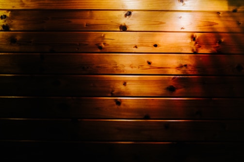 Wooden planks with a rich, dark finish are illuminated by a warm, soft light. The surface has a natural texture with visible knots and grain patterns, adding character to the wood.