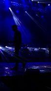 A musician is silhouetted against a dark stage with bright blue lights beaming from above and behind. The beams create a dynamic and dramatic atmosphere, highlighting the musician's posture with an instrument in hand while a drum set is faintly visible in the background.