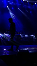 A musician is silhouetted against a dark stage with bright blue lights beaming from above and behind. The beams create a dynamic and dramatic atmosphere, highlighting the musician's posture with an instrument in hand while a drum set is faintly visible in the background.
