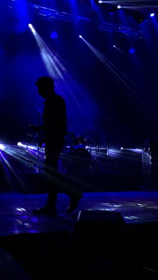 A musician is silhouetted against a dark stage with bright blue lights beaming from above and behind. The beams create a dynamic and dramatic atmosphere, highlighting the musician's posture with an instrument in hand while a drum set is faintly visible in the background.