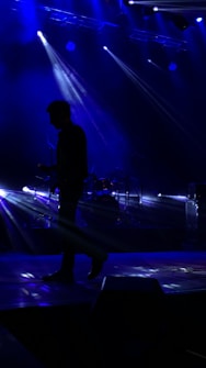 A musician is silhouetted against a dark stage with bright blue lights beaming from above and behind. The beams create a dynamic and dramatic atmosphere, highlighting the musician's posture with an instrument in hand while a drum set is faintly visible in the background.