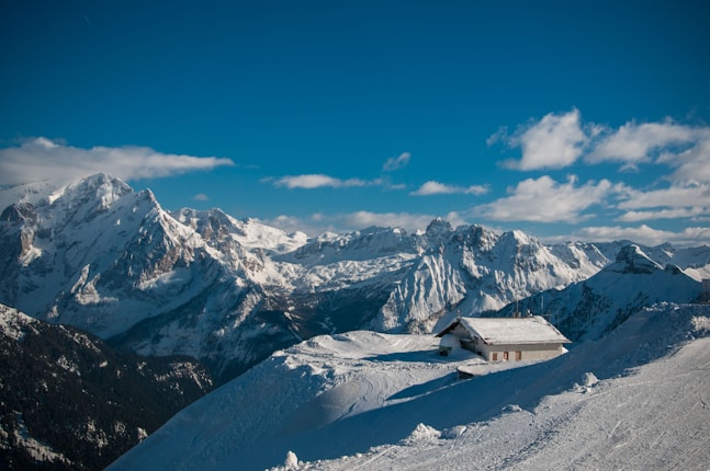 A snow-covered mountain range under clear blue skies, with a small cabin or building situated in the foreground on a snow-blanketed slope.