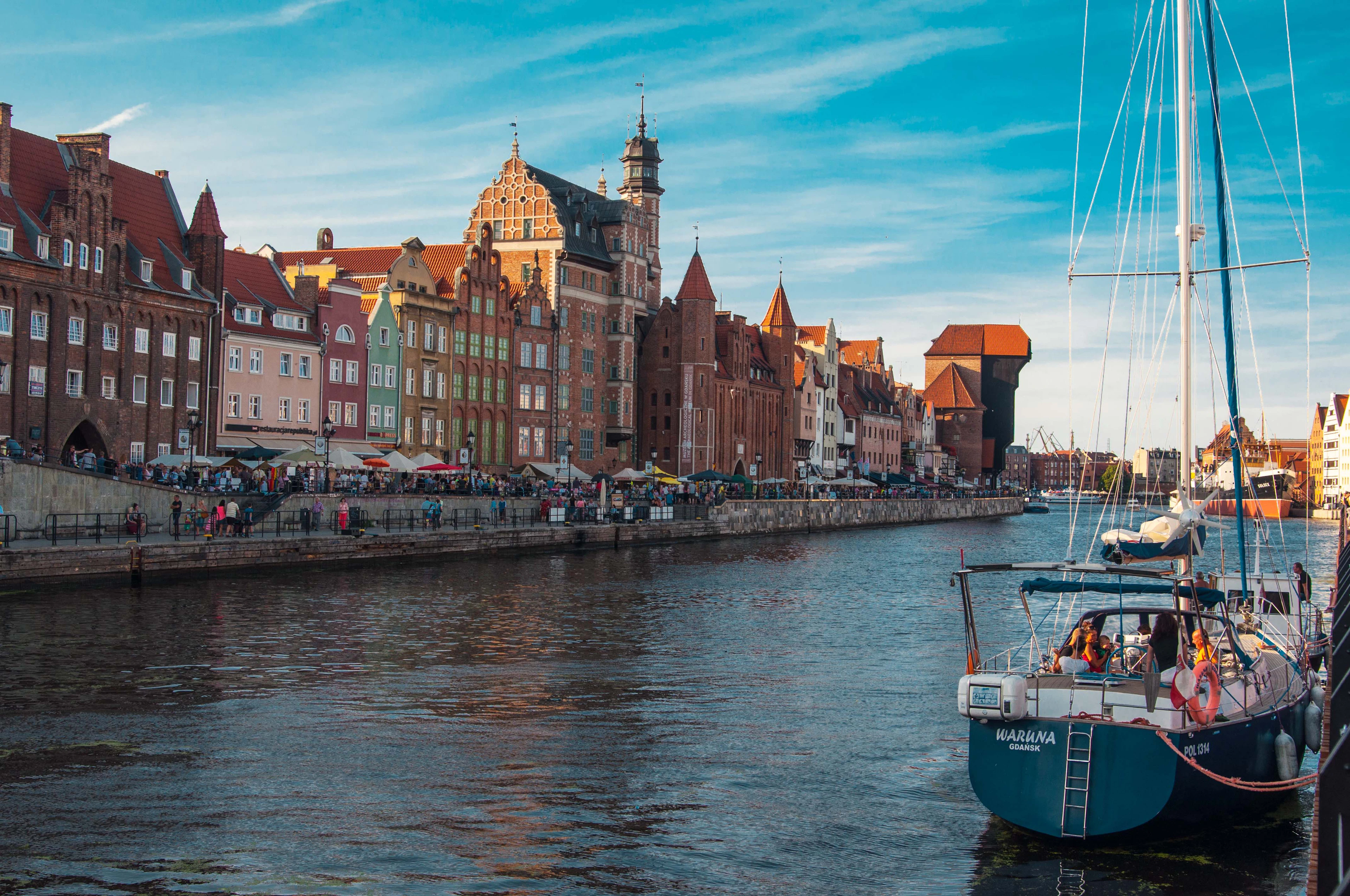 A sailboat's mast frames Gdańsk's Motława River at golden hour, with the Crane tower and colorful embankment houses beyond.