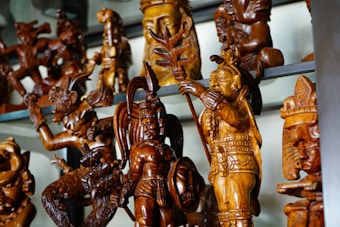 A collection of intricately carved wooden figures displayed on a shelf, featuring detailed human and mythical forms with elaborate headdresses and expressions. The craftsmanship highlights rich textures and patterns in the wood.
