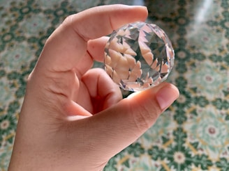 A hand is holding a clear, faceted crystal sphere. The background is a detailed, patterned green and yellow surface, likely a decorative tile or fabric. The lighting causes the crystal to create reflections and light patterns on the hand and surrounding area.