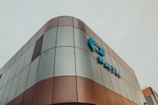 Modern architectural building with a curved exterior, featuring a metallic surface with a mix of bronze and grey panels. Blue signage is prominently displayed on the facade.