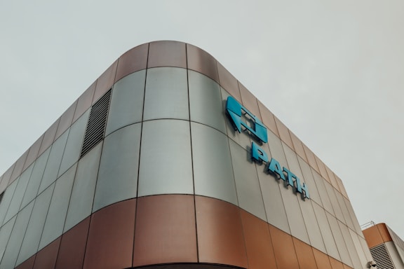 Modern architectural building with a curved exterior, featuring a metallic surface with a mix of bronze and grey panels. Blue signage is prominently displayed on the facade.