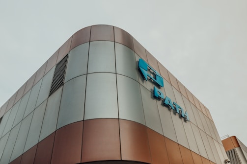 Modern architectural building with a curved exterior, featuring a metallic surface with a mix of bronze and grey panels. Blue signage is prominently displayed on the facade.