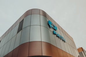 Modern architectural building with a curved exterior, featuring a metallic surface with a mix of bronze and grey panels. Blue signage is prominently displayed on the facade.