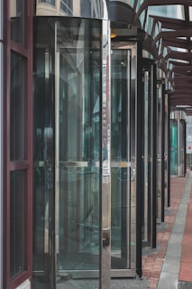 Several glass revolving doors are arranged in a row along the exterior of a building. The doors are framed by metal and are designed to allow entry and exit. The structure of the building is also visible with large windows and a covered walkway leading up to the doors.
