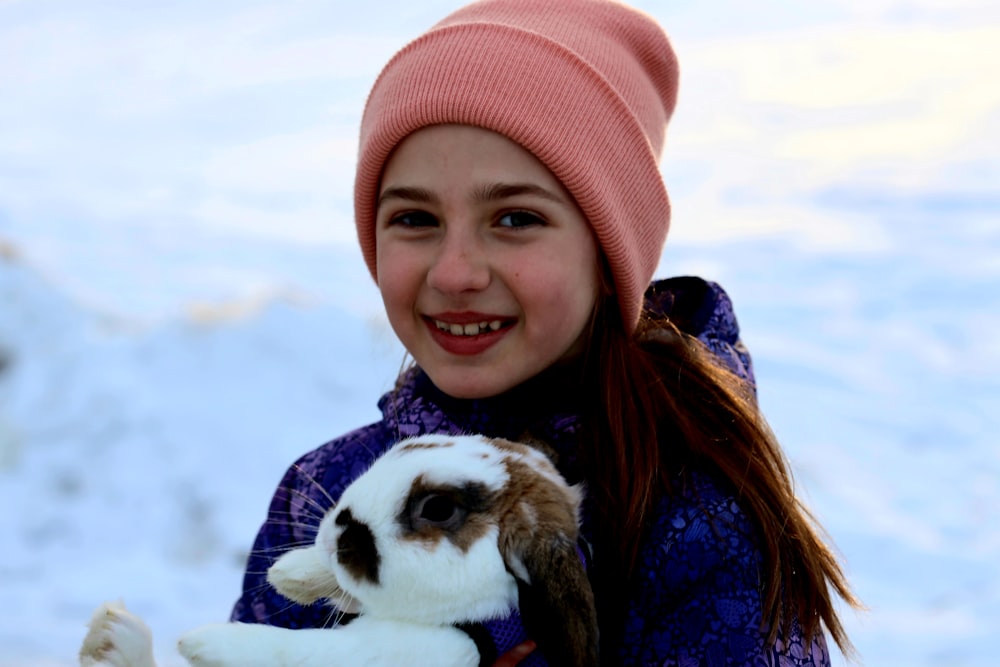 girl in blue jacket and pink knit cap holding white and brown short coated dog during