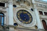 A detailed view of an ornate astronomical clock featuring zodiac symbols in gold on a deep blue background. The clock is set in a historic building facade with Roman numerals surrounding the circumference.