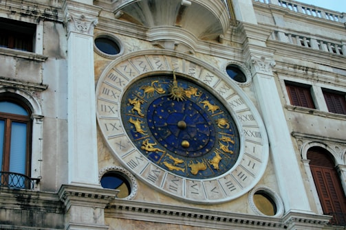 A detailed view of an ornate astronomical clock featuring zodiac symbols in gold on a deep blue background. The clock is set in a historic building facade with Roman numerals surrounding the circumference.