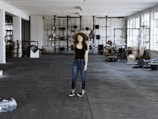 A person wearing a black shirt, blue jeans, and a brown hat stands in the middle of a spacious and industrial-style warehouse. The warehouse is filled with various equipment and items scattered around, such as cables, metal structures, and machinery parts. Large windows on the right allow natural light to stream in, casting shadows on the concrete floor.