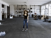 A person wearing a black shirt, blue jeans, and a brown hat stands in the middle of a spacious and industrial-style warehouse. The warehouse is filled with various equipment and items scattered around, such as cables, metal structures, and machinery parts. Large windows on the right allow natural light to stream in, casting shadows on the concrete floor.