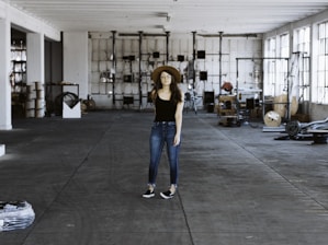 A person wearing a black shirt, blue jeans, and a brown hat stands in the middle of a spacious and industrial-style warehouse. The warehouse is filled with various equipment and items scattered around, such as cables, metal structures, and machinery parts. Large windows on the right allow natural light to stream in, casting shadows on the concrete floor.