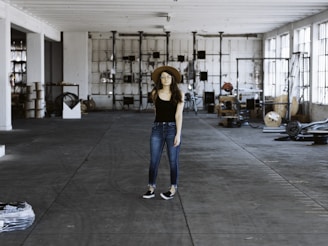 A person wearing a black shirt, blue jeans, and a brown hat stands in the middle of a spacious and industrial-style warehouse. The warehouse is filled with various equipment and items scattered around, such as cables, metal structures, and machinery parts. Large windows on the right allow natural light to stream in, casting shadows on the concrete floor.