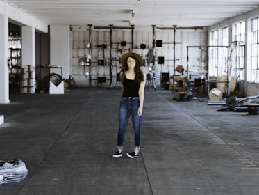 A person wearing a black shirt, blue jeans, and a brown hat stands in the middle of a spacious and industrial-style warehouse. The warehouse is filled with various equipment and items scattered around, such as cables, metal structures, and machinery parts. Large windows on the right allow natural light to stream in, casting shadows on the concrete floor.