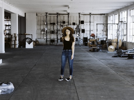 A person wearing a black shirt, blue jeans, and a brown hat stands in the middle of a spacious and industrial-style warehouse. The warehouse is filled with various equipment and items scattered around, such as cables, metal structures, and machinery parts. Large windows on the right allow natural light to stream in, casting shadows on the concrete floor.