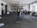 A person wearing a black shirt, blue jeans, and a brown hat stands in the middle of a spacious and industrial-style warehouse. The warehouse is filled with various equipment and items scattered around, such as cables, metal structures, and machinery parts. Large windows on the right allow natural light to stream in, casting shadows on the concrete floor.