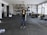 A person wearing a black shirt, blue jeans, and a brown hat stands in the middle of a spacious and industrial-style warehouse. The warehouse is filled with various equipment and items scattered around, such as cables, metal structures, and machinery parts. Large windows on the right allow natural light to stream in, casting shadows on the concrete floor.