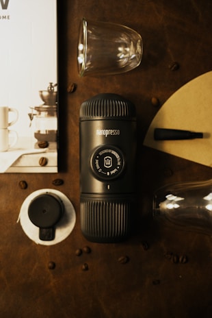 A compact black Nanopresso coffee maker is positioned on a dark brown leather surface surrounded by scattered coffee beans. Nearby, there's a pair of transparent espresso glasses, a circular black coffee tamper, and a small black pen-like accessory. In the background, a partial view of a booklet or magazine featuring a coffee setup is visible.