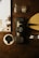 A compact black Nanopresso coffee maker is positioned on a dark brown leather surface surrounded by scattered coffee beans. Nearby, there's a pair of transparent espresso glasses, a circular black coffee tamper, and a small black pen-like accessory. In the background, a partial view of a booklet or magazine featuring a coffee setup is visible.