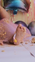 A close-up of a baby's foot covered in pastel-colored cake and frosting. The baby is wearing a pink tulle skirt, and pieces of cake are scattered on the white floor. In the background, colorful balloons and streamers create a festive atmosphere.