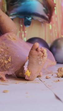A close-up of a baby's foot covered in pastel-colored cake and frosting. The baby is wearing a pink tulle skirt, and pieces of cake are scattered on the white floor. In the background, colorful balloons and streamers create a festive atmosphere.