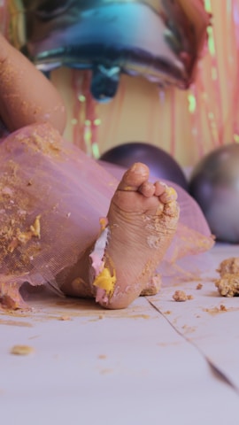 A close-up of a baby's foot covered in pastel-colored cake and frosting. The baby is wearing a pink tulle skirt, and pieces of cake are scattered on the white floor. In the background, colorful balloons and streamers create a festive atmosphere.