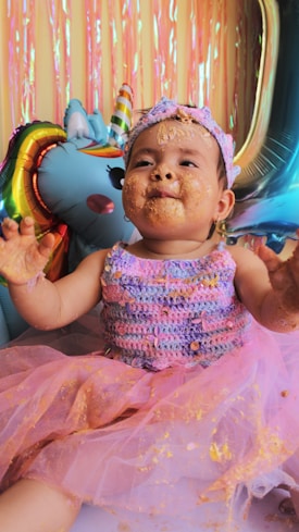 A baby in a colorful outfit, covered in cake, is sitting happily next to a large unicorn balloon. The background features shiny, colorful streamers.