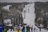 A snow-covered ski resort with a busy ski lift in the center and numerous people dressed in winter gear gathered at the base. The slope is surrounded by tall evergreen trees, and several skiers are visible on the hill. The sky is overcast, contributing to a wintry atmosphere.