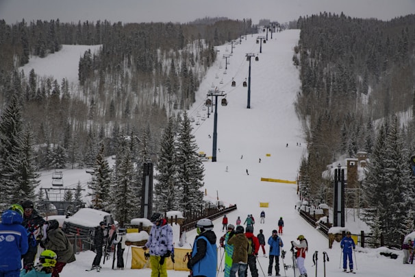 A snow-covered ski resort with a busy ski lift in the center and numerous people dressed in winter gear gathered at the base. The slope is surrounded by tall evergreen trees, and several skiers are visible on the hill. The sky is overcast, contributing to a wintry atmosphere.