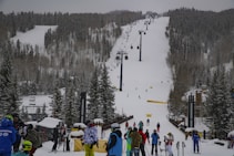 A snow-covered ski resort with a busy ski lift in the center and numerous people dressed in winter gear gathered at the base. The slope is surrounded by tall evergreen trees, and several skiers are visible on the hill. The sky is overcast, contributing to a wintry atmosphere.